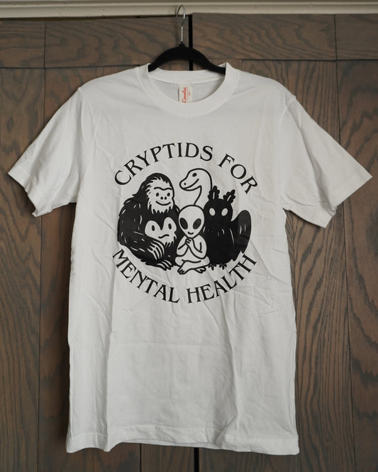 White t-shirt with black graphic design featuring bigfoot, mothman, nessie and an alien, with the words cryptids for mental health