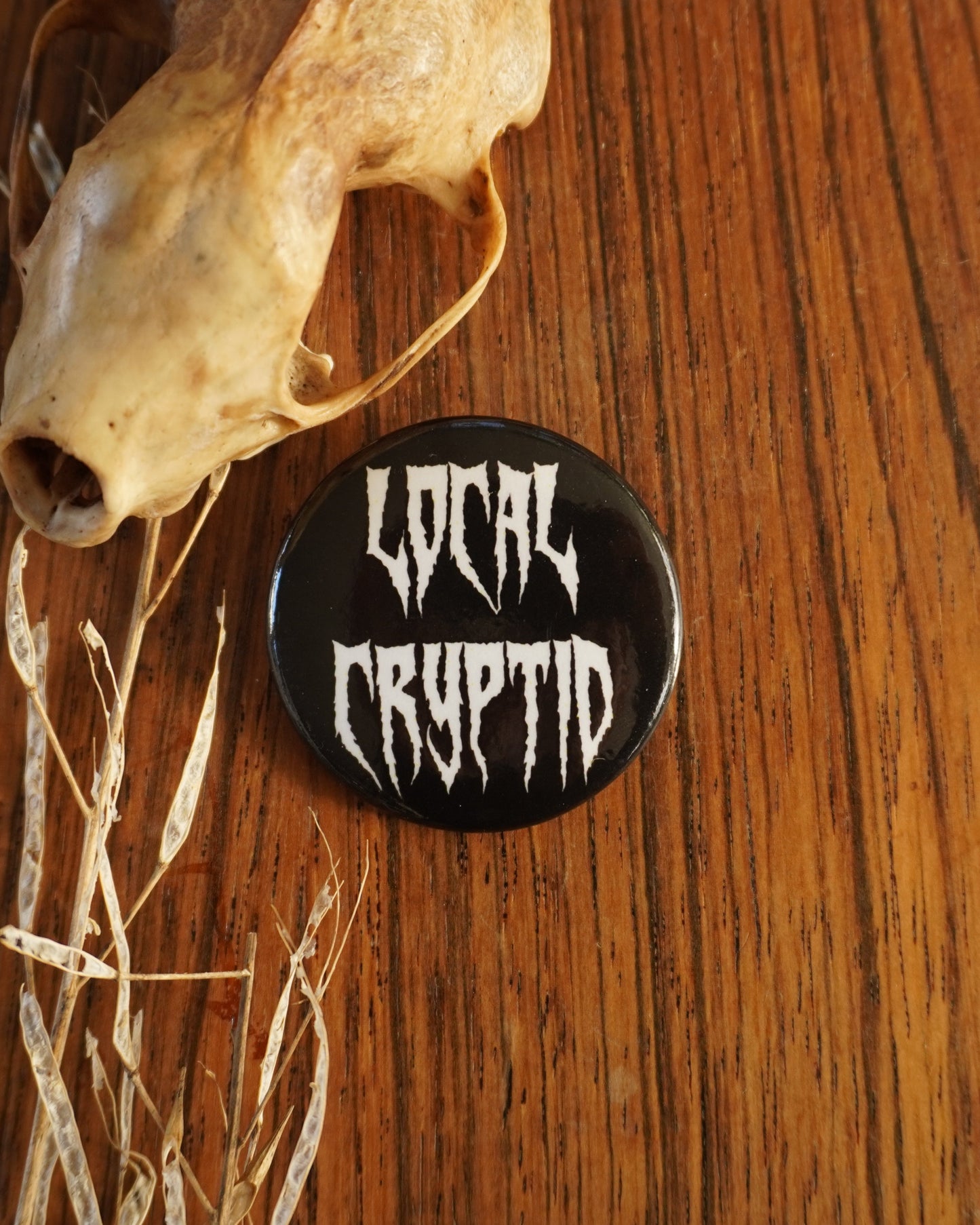 Button with 'Local Cryptid' text on a wooden surface with a skull and grass.