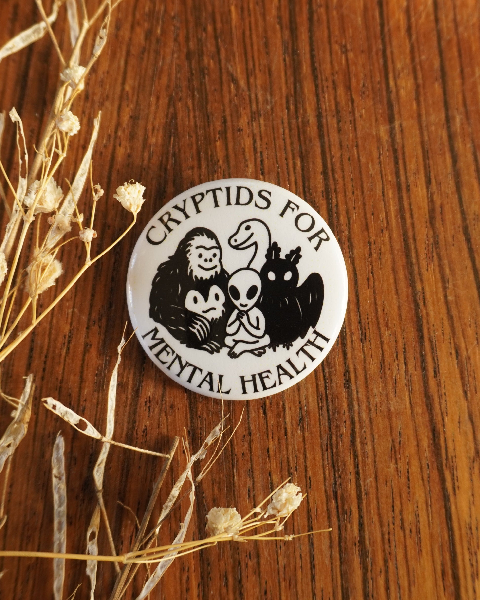 Round badge with 'Cryptids for Mental Health' design on a wooden surface with dried plants.