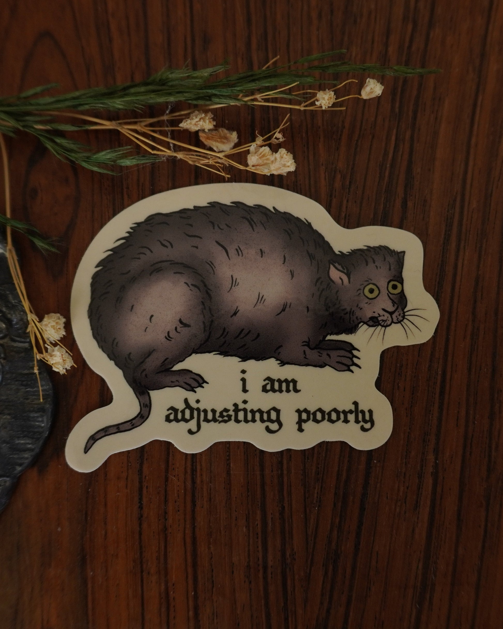 Sticker of a cat with text 'I am adjusting poorly' on a wooden surface with a candle and flowers.