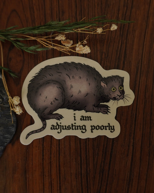 Sticker of a cat with text 'I am adjusting poorly' on a wooden surface with a candle and flowers.