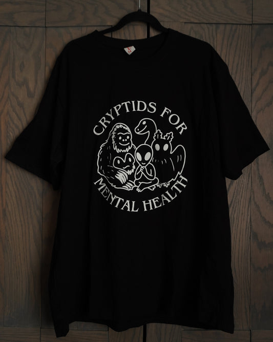 Black t-shirt with a white graphic design featuring bigfoot, an alien, mothman and nessie, it says cryptids for mental health