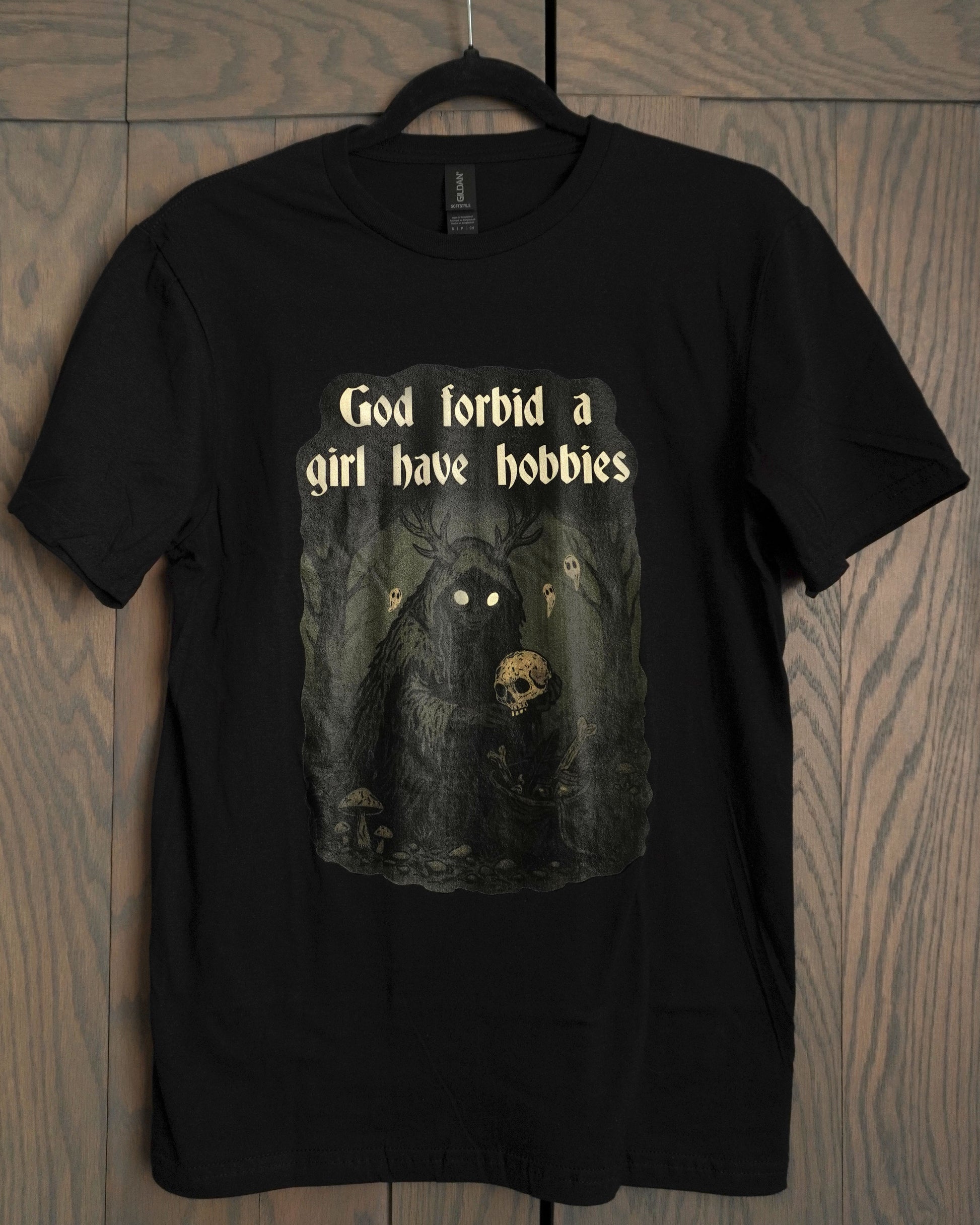 a black tshirt with a dark green scene with a hooded gremlin collecting bones in the forest, with the words god forbid a girl have hobbies