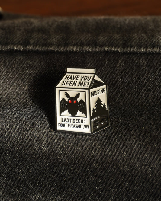 Enamel pin of a milk carton with mothman and text on a dark fabric background. It says have you seen me? Last seen: Point Pleasant WV